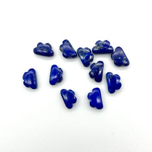 Pode incluir: Uma coleção de pequenas contas de lápis-lazúli em forma de nuvem. As contas são de cor azul profundo com manchas douradas e estão espalhadas sobre uma superfície branca. Estas contas são provavelmente usadas para fazer joias ou outros projetos de artesanato.