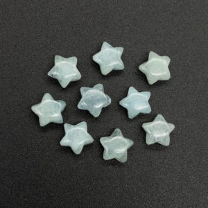 Könnte beinhalten: Neun hellblaue, sternförmige Edelsteine liegen auf einer dunkelgrauen Oberfläche. Die Edelsteine sind durchscheinend, mit sichtbaren inneren Mustern. Jeder Stern hat fünf Zacken und eine leicht abgerundete Form.