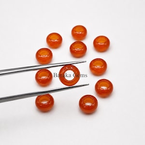 May include: A collection of eleven round, smooth, translucent orange carnelian beads. One bead is held by silver-toned metal tweezers, displaying a central hole. The text Baraka Gems is printed in black in the center of the arrangement of beads.