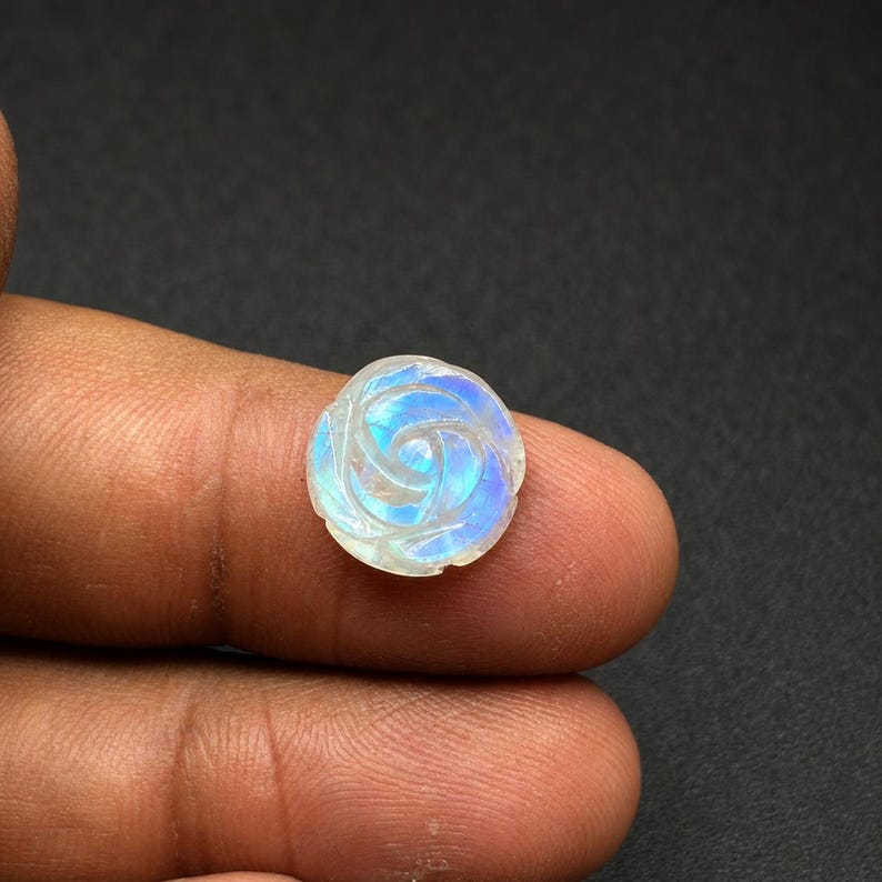 Puede incluir: Una gema tallada en forma de rosa. La piedra muestra colores iridiscentes, incluyendo azul, blanco y toques de p&uacute;rpura. El tallado de la rosa es detallado, con p&eacute;talos visibles.