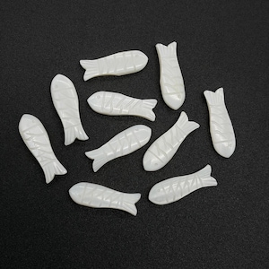 Może przedstawiać: Kolekcja jedenastu białych koralików w kształcie ryb, każdy z rzeźbionymi detalami. Koraliki są rozrzucone na ciemnej powierzchni. Ryby różnią się kształtem, niektóre są wydłużone, inne bardziej kompaktowe, wszystkie o teksturowanym wyglądzie.