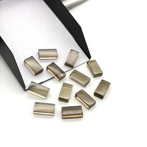 May include: Rectangular smoky quartz gemstones, scattered on a white surface. The translucent, brownish-grey gemstones are polished. They are next to a black and white container. The gemstones are likely for jewellery making.