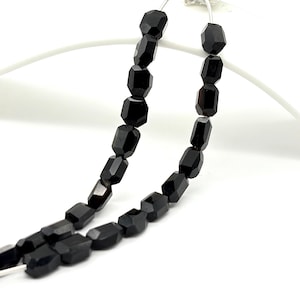 May include: Two strands of black gemstone beads. Each bead is a faceted, rectangular shape, strung together with a thin, silver-coloured wire. The beads have a polished, reflective surface, and are set against a white background.