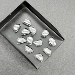 Könnte beinhalten: Eine Sammlung von elf kleinen, klaren, wolkenförmigen Glaskabochons, die auf einem schwarzen Metalltablett angeordnet sind. Jedes Stück hat eine glatte, abgerundete Oberfläche mit einem durchscheinenden Aussehen, das Licht auf den polierten Kanten reflektiert.