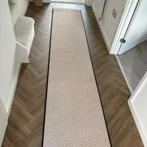May include: A beige runner rug with a black border, laid on a herringbone wooden floor. The rug features a chevron pattern. The hallway is bright, with white walls and a glimpse of a doorway. The rug is suitable for hallways.