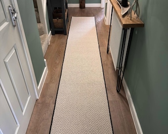 May include: A long, rectangular beige rug with a black border, featuring a chevron pattern. The rug is placed in a hallway with wooden flooring and green walls. The hallway also includes a console table and decorative items.