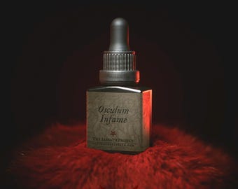 May include: A small glass bottle with a black dropper top. The bottle is labelled "Osculum Infame" and "The Sabbath Project" with a red star and website address. The bottle is sitting on a red fluffy surface.