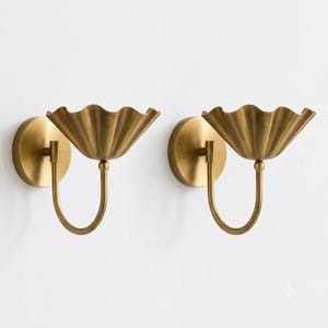 May include: A pair of wall sconces featuring a brushed brass finish. Each fixture consists of a circular wall-mounted plate, a curved arm, and a wide, scalloped, bowl-shaped shade that flares outward at the top edge in a petal-like design.