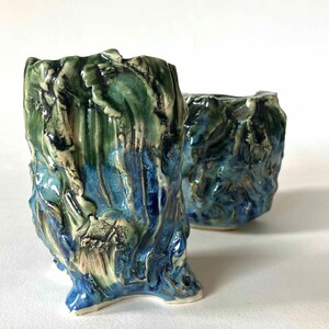 May include: Two ceramic vases with a textured, organic design. The vases feature a colour palette of blues and greens, with a glossy finish. The larger vase has a unique, three-footed base. The smaller vase is in the background.