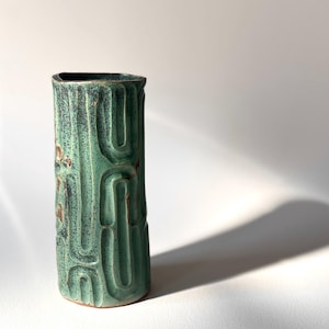 May include: A tall, cylindrical ceramic vase with a textured, carved design. The vase is primarily a mottled teal green colour with darker green and brown accents. The vase has a rectangular opening at the top and is set against a white background.