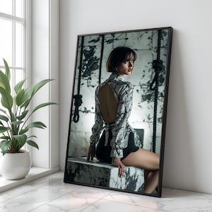 May include: A framed art print featuring a person in a patterned top and black shorts. The artwork has a black frame and is leaning against a white wall. The person is sitting on a stone block, with ropes hanging in the background.