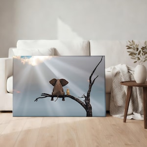 May include: A canvas print depicts an elephant and a bird perched on a tree branch, gazing at a bright sky with sun rays. The elephant is brown, and the bird is yellow. The artwork is displayed in a living room setting.