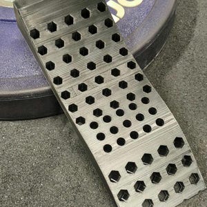 以下が含まれることがあります： 黒色のウェイトリフティングウェッジ。表面はテクスチャ加工され、六角形と円形の切り欠きが並んでいます。ウェッジは角度がついており、エクササイズ中に踵を持ち上げるように設計されています。背景にはウェイトプレートが含まれています。