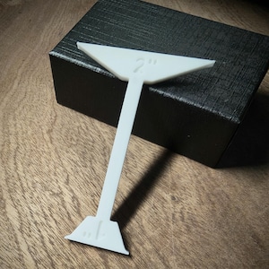 May include: A white plastic measuring tool with a triangular top and a long, thin handle. The top of the tool is labeled with the number "2". The tool is designed for measuring and is placed on a black textured surface.