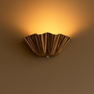 May include: A brass-colored wall sconce with a scalloped, fan-like design. The light fixture is mounted on a neutral-toned wall, casting a warm glow. The sconce has a small, round decorative element at the bottom.
