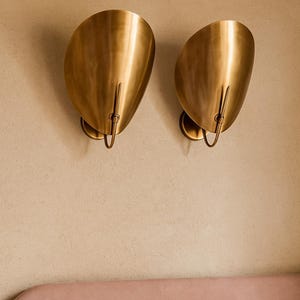 May include: Two gold-colored wall sconces with a curved, shell-like design. Each sconce has a small, circular base and a curved metal piece extending downwards. The sconces are mounted on a beige wall above a pink headboard and white pillows.