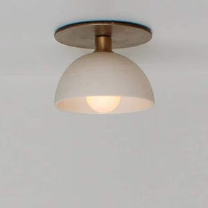 May include: A modern ceiling light fixture with a white, dome-shaped shade and a brass-colored base. The light is illuminated, casting a warm glow. The fixture is designed for indoor use.