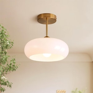 Op de afbeelding: Een moderne plafondlamp met een witte, ovaalvormige glazen kap. Het licht hangt aan een messingkleurige steel en een cirkelvormige plafondbevestiging. De armatuur is ontworpen voor sfeerverlichting.
