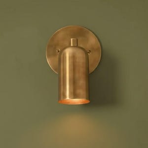 May include: A brass wall sconce with a cylindrical light fixture and a circular backplate. The sconce is mounted on a green wall. The light fixture is illuminated, casting a warm glow.