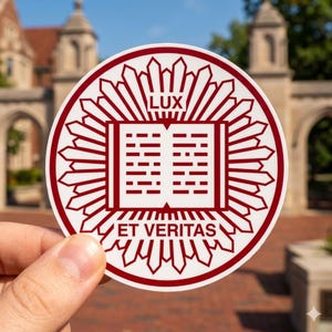 May include: A round white sticker with a maroon emblem. The emblem features an open book with the words "LUX" above and "ET VERITAS" below. The sticker is held in front of a blurred background of a building.