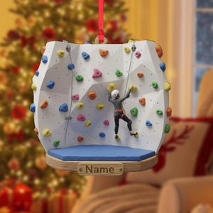 May include: A white climbing wall ornament with colorful holds and a figure scaling it. The ornament has a blue base and a wooden nameplate. A red ribbon is attached for hanging. The background includes a Christmas tree.