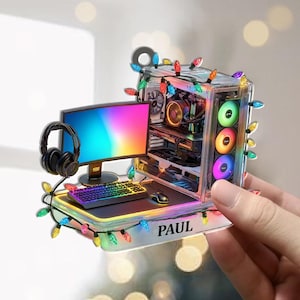 May include: A clear acrylic ornament shaped like a gaming PC setup, complete with a monitor, keyboard, mouse, and headphones. The PC case has colorful LED lights and is adorned with Christmas lights. The name "PAUL" is printed on the base.