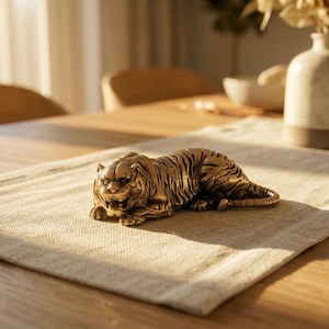 Könnte beinhalten: Eine bronzefarbene Tigerfigur in Ruheposition. Die detaillierte Skulptur weist Streifen und einen grimmigen Ausdruck auf. Sie liegt auf einem beigen Leinen-Tischläufer, mit einem Holztisch und einer Vase im Hintergrund. Die Figur ist etwa 15 cm lang.