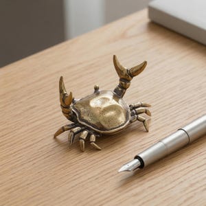 May include: A detailed brass crab figurine with raised claws and legs. The crab is positioned on a light wood surface next to a silver fountain pen. The crab's body has a textured surface.