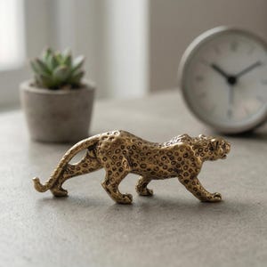 Può includere: Una statuetta di leopardo dorato con una superficie strutturata e un motivo maculato. La scultura decorativa è in posizione di camminata. Una piccola pianta succulenta in un vaso di cemento e un orologio bianco sono sullo sfondo.