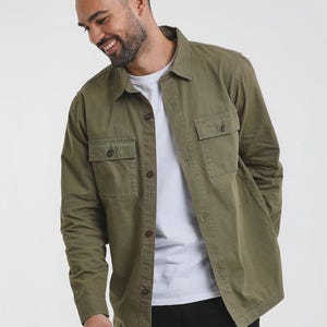 May include: Olive green long-sleeve shirt with two chest pockets and button closures. The shirt is worn over a white t-shirt. The model is smiling and wearing black pants. The shirt is a casual, everyday garment.