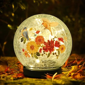 May include: A decorative glass globe with a crackled finish, featuring illustrations of birds and colorful flowers. The globe is illuminated with small lights and sits on a black base, creating a warm, inviting glow. The overall theme is nature and home decor.