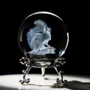 May include: A clear glass sphere with a detailed 3D engraving of a squirrel perched on a branch. The sphere sits on an ornate silver-colored metal stand with curved legs. The background is black.