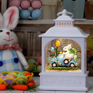May include: White Easter-themed lantern with a decorative scene inside. The scene features a blue truck carrying a bunny, eggs, and a sign that says "Happy Easter." The lantern has a handle and decorative accents.