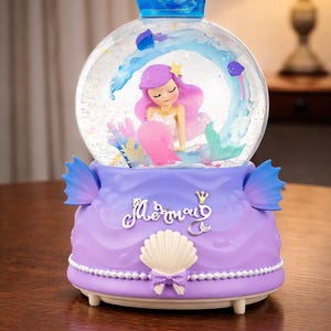 May include: A decorative snow globe featuring a mermaid figure with purple hair and a pink tail inside a glass globe. The base is purple with the word "Mermaid" in script. A blue crown tops the globe.