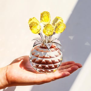 May include: A decorative crystal vase with yellow rose flowers. The vase is clear and faceted, with a round shape. The roses are a bright yellow color, and the stems are silver. The vase is held in a hand.