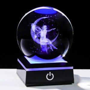 May include: A clear crystal ball with a 3D image of a fairy sitting on a crescent moon, illuminated with blue light. The sphere sits on a black base with a power button, creating a magical, decorative piece.