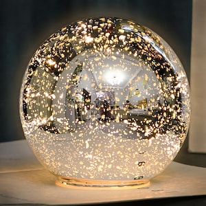 May include: A spherical, silver-colored decorative lamp with a speckled pattern of warm, golden-yellow lights. The lamp reflects the surrounding environment, creating a mirrored effect. The lamp sits on a light-colored surface.
