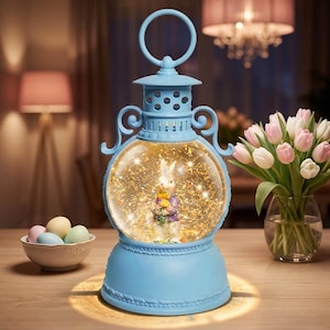 May include: A light blue Easter-themed snow globe lantern with a bunny figurine inside. The globe is surrounded by gold glitter and small lights. A bouquet of pink and white tulips and a bowl of pastel eggs are also visible.