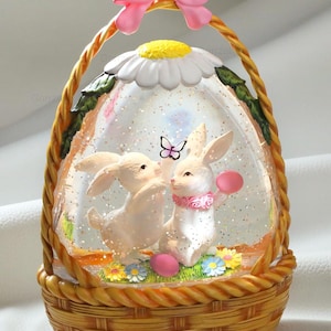 May include: A decorative Easter-themed snow globe in a woven basket design. Inside, two white bunnies are depicted, along with a butterfly and pink eggs. The globe is topped with a daisy and a pink bow.