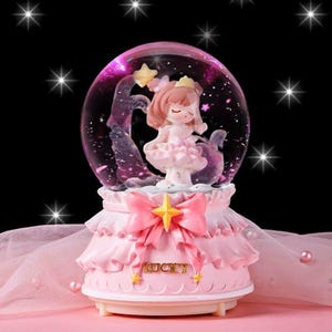 May include: A pink snow globe featuring a small figurine of a girl in a dress, with a pink bow and the word "LUCKY". The globe has a pink and purple background with stars. The base is pink with a ruffled design.