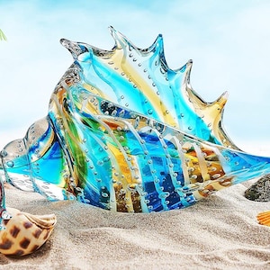 May include: Three colorful glass sculptures of sea creatures: a fish, a seashell, and an octopus. The fish is blue, orange, and yellow. The seashell is blue, yellow, and clear. The octopus is orange and clear. They are on a sandy surface.
