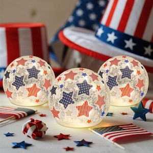 May include: Three illuminated glass globes decorated with red, white, and blue stars. The globes are surrounded by patriotic decorations, including an Uncle Sam hat and small American flags. The overall theme is a Fourth of July or patriotic celebration.