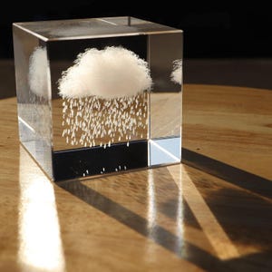 May include: A clear glass cube with a fluffy white cloud and falling rain design. The cube sits on a wooden surface, casting a shadow. The cloud and rain are white, creating a whimsical, artistic piece.
