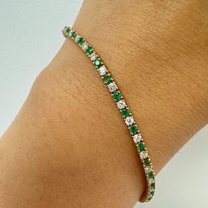Puede incluir: Una pulsera dorada con piedras preciosas de esmeraldas y diamantes alternas. La pulsera está engastada con pequeñas piedras redondas, creando una línea continua de color y brillo. La pulsera se lleva en la muñeca.