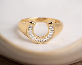 Könnte beinhalten: Goldfarbener Ring mit Hufeisen-Design. Das Hufeisen ist mit kleinen, rechteckigen, klaren Steinen besetzt. Der Ring liegt auf einer weißen Oberfläche.
