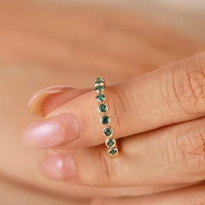 Può includere: Un anello d'oro con una serie di pietre preziose rotonde verde smeraldo. L'anello è incastonato in una fascia sottile, con le pietre equidistanti. È un gioiello delicato.
