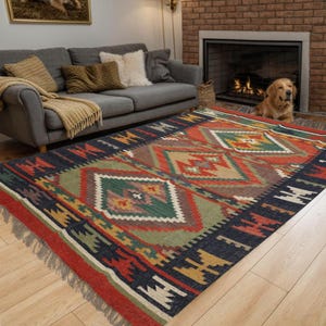 May include: A colorful area rug with a geometric pattern in shades of red, green, blue, and gold. The rug has a fringed edge and is placed on a hardwood floor. A gray sofa and a fireplace are in the background.