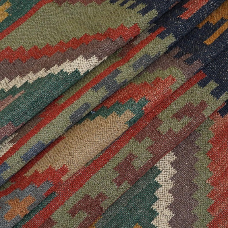 May include: A folded, woven rug with a geometric pattern. The rug features a mix of colors, including green, red, brown, and blue. The design includes diagonal stripes and zig-zag patterns, creating a textured appearance.