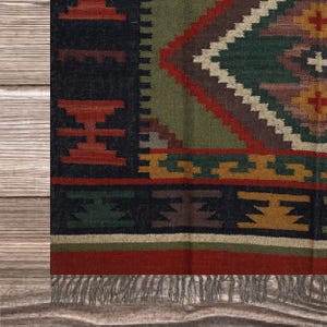May include: Close-up of a colorful woven rug with a geometric pattern. The rug features a black border with red and green accents, and a central diamond design in shades of green, brown, and cream. The rug has a fringed edge.