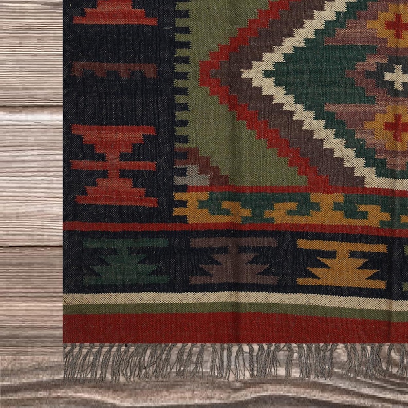 May include: Close-up of a colorful woven rug with a geometric pattern. The rug features a black border with red and green accents, and a central diamond design in shades of green, brown, and cream. The rug has a fringed edge.